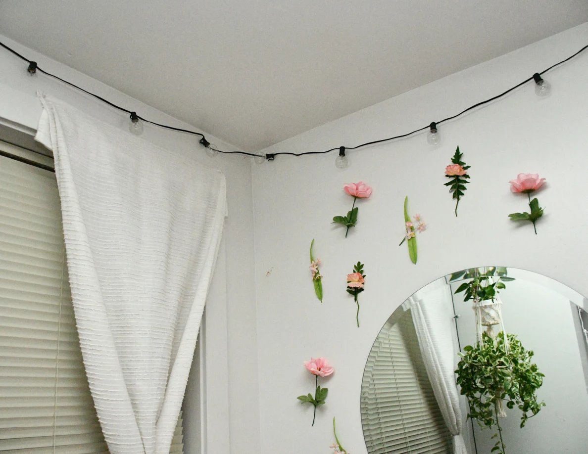 String lights above flower-decorated wall, round mirror and curtained window.