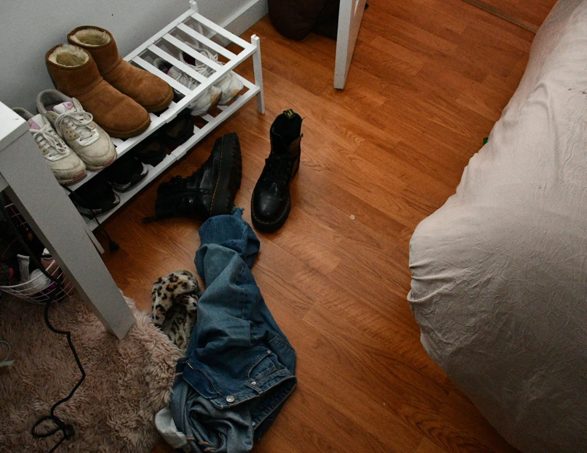 Black boots and jeans on wood floor beside shoe rack holding tan boots and sneakers.