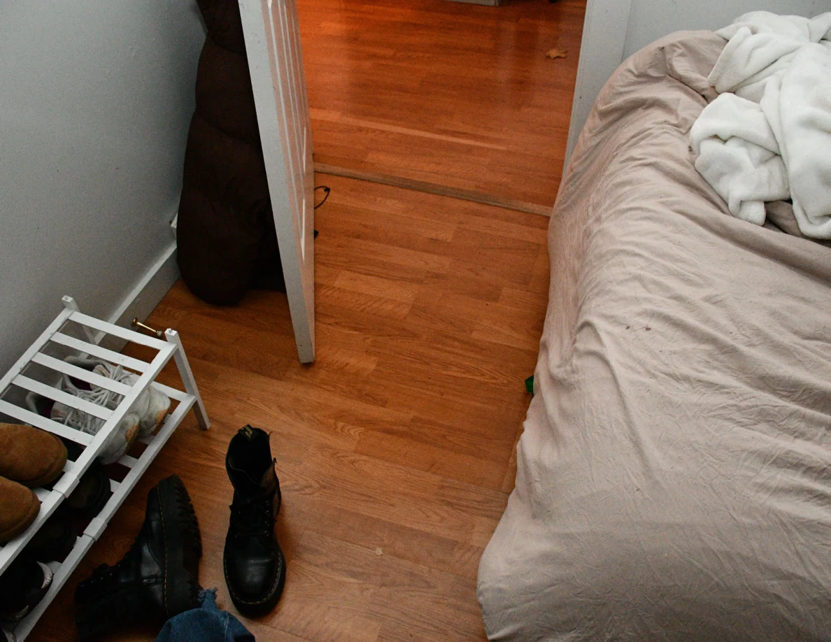 Wood floor between bed and open doorway, with shoe rack and black boot.