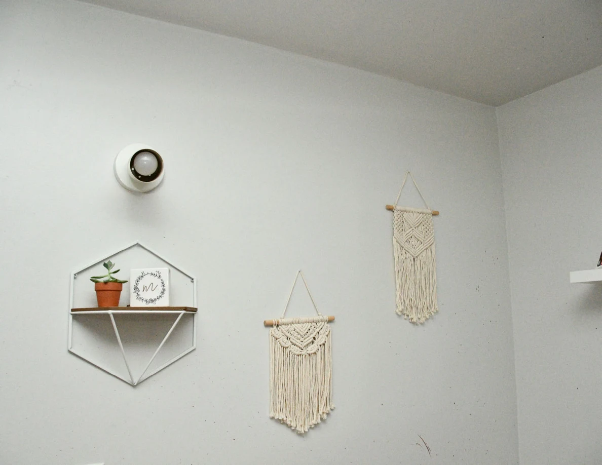 White wall light above geometric plant shelf and two woven wall hangings.