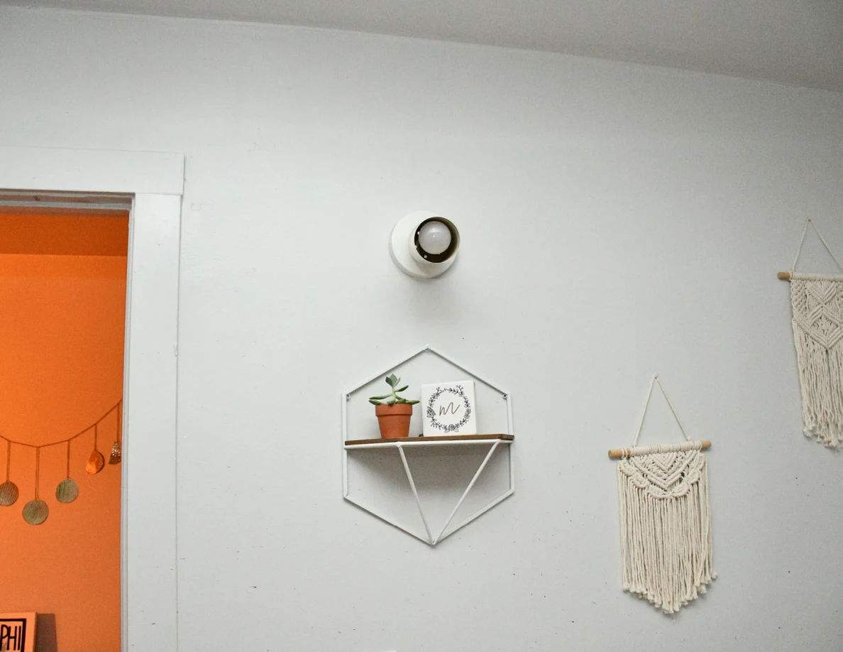 Wall light, geometric plant shelf and woven wall hanging beside open doorway.