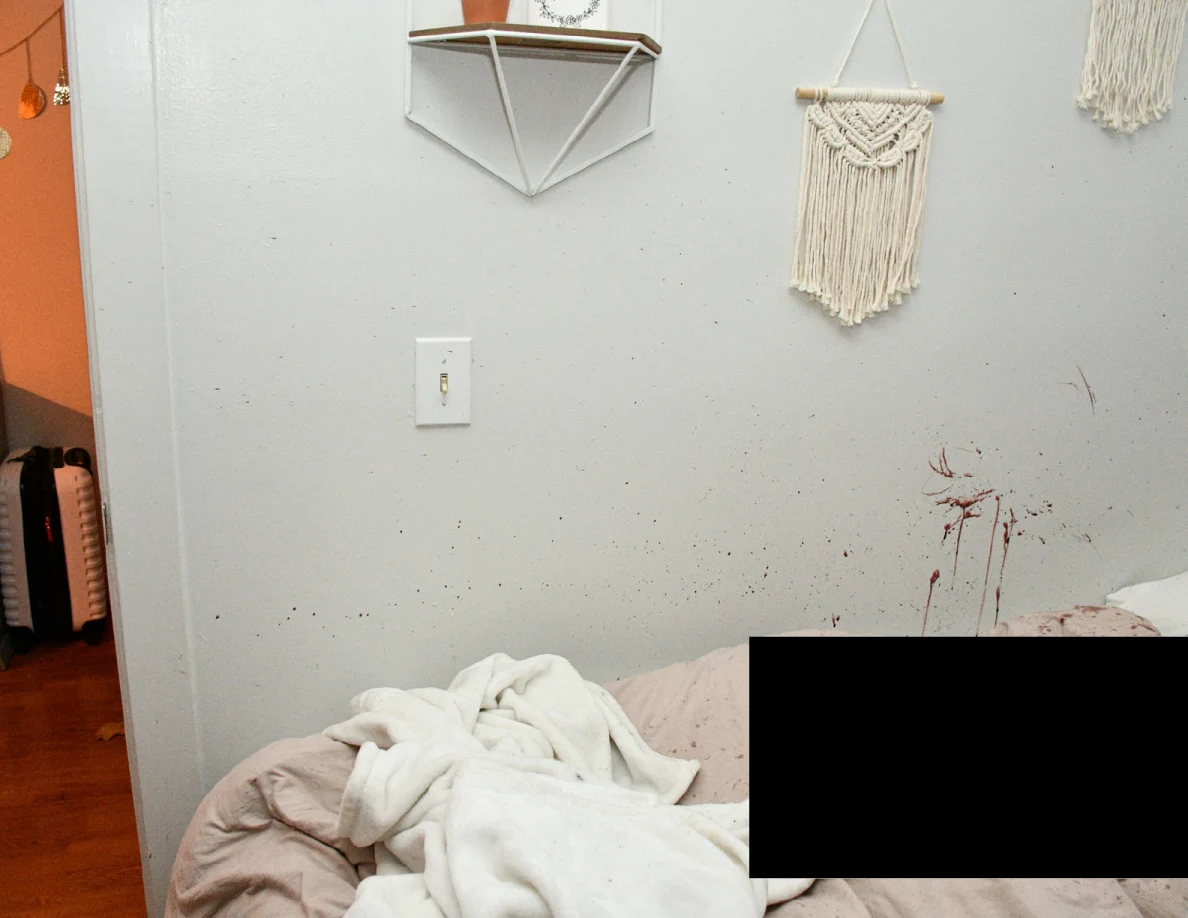 Wall shelf and woven hanging above reddish wall marks and rumpled white blanket, with a black redaction.