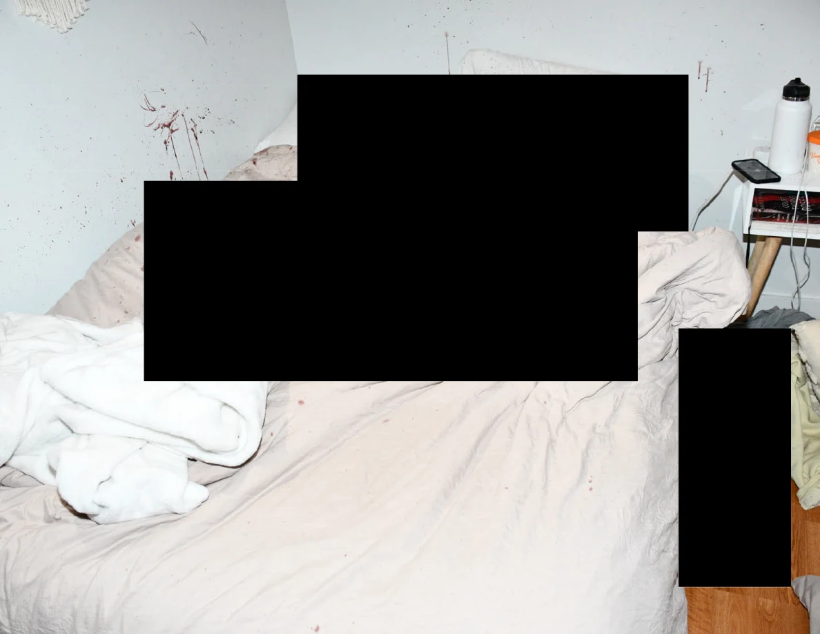 Bright view of bed, bedside table and reddish wall marks around black redactions.