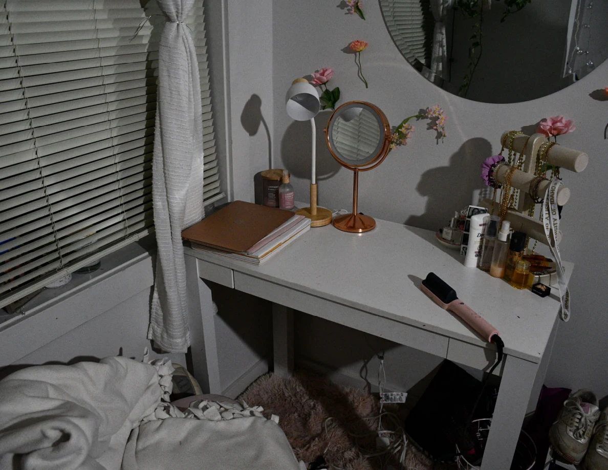 White desk with closed laptop, mirrors, jewelry stand, cosmetics and hair-styling tool beside a curtained window.