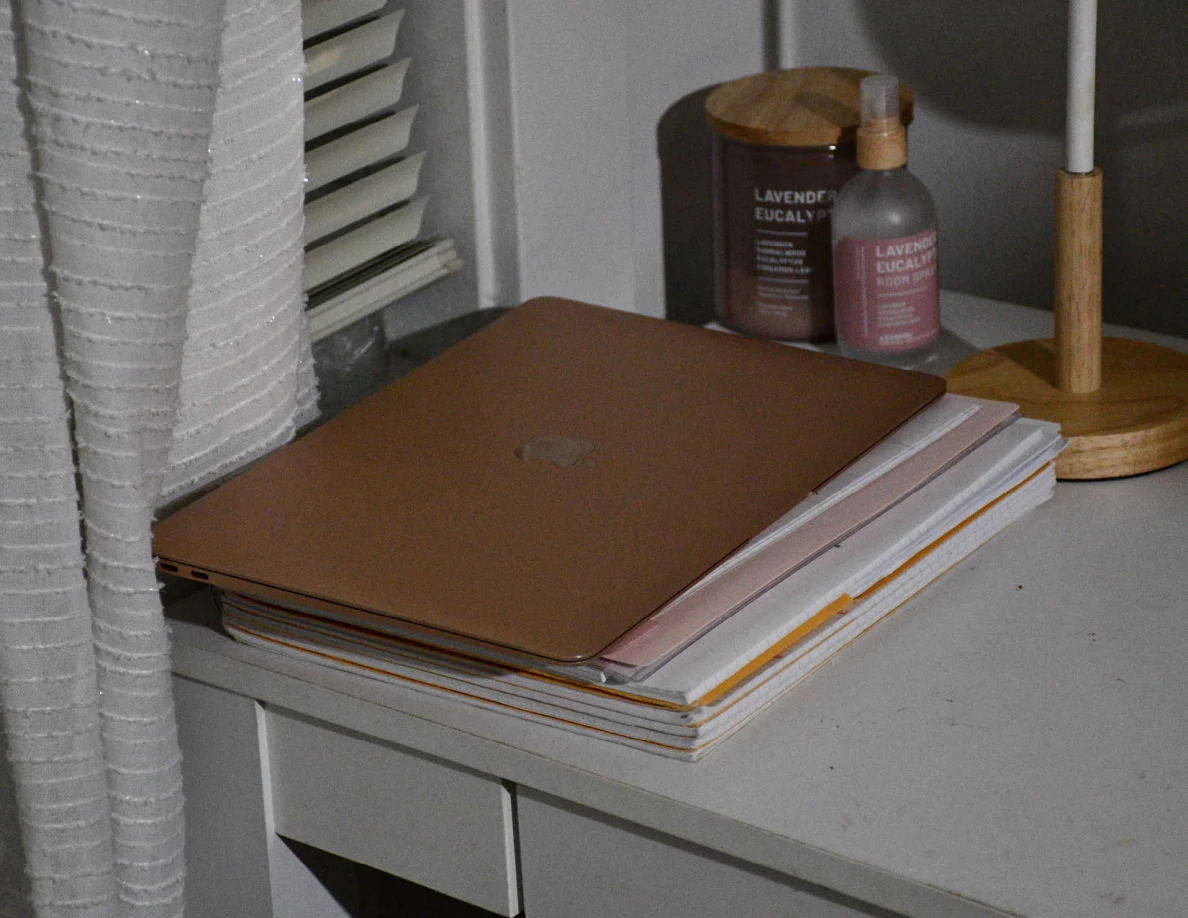 Closed rose-colored laptop on stacked papers beside candle, spray bottle and desk lamp.