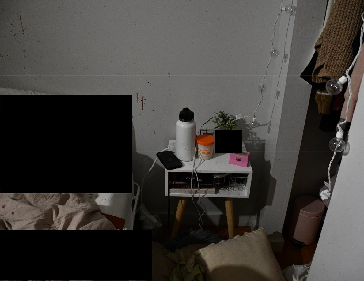 Small bedside table with phone and white bottle beside open closet and redacted bedding.