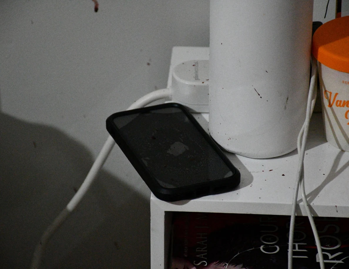Black phone beside white bottle, orange-lidded container and charging cables on bedside table.