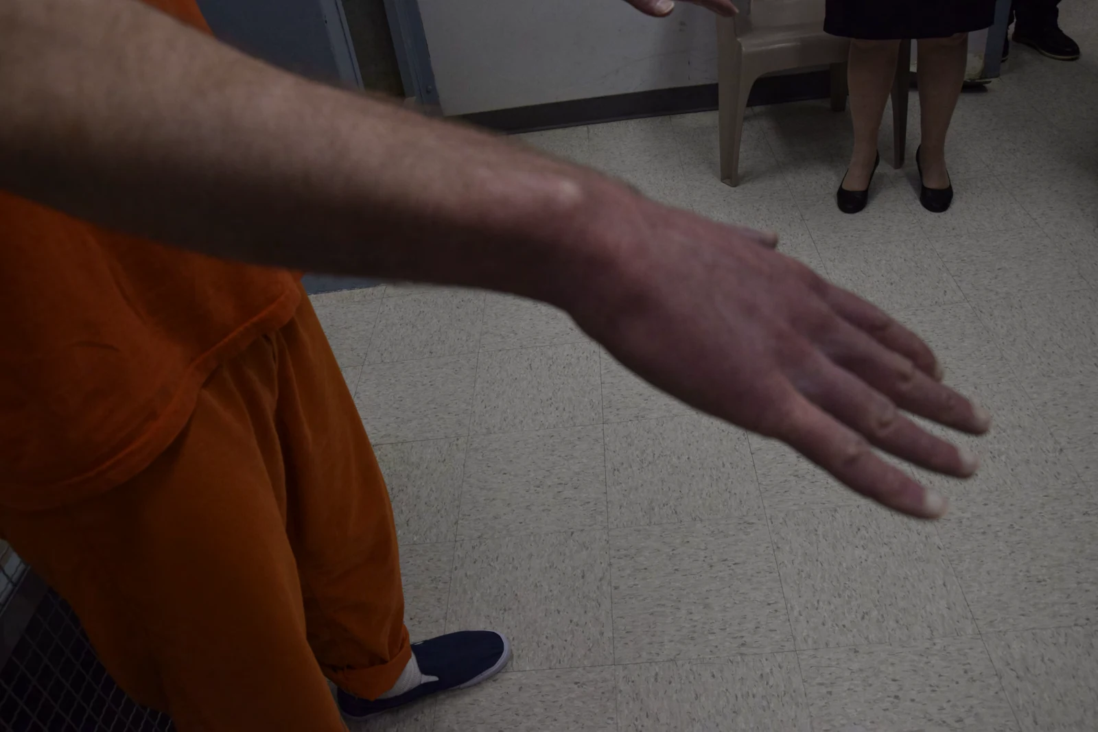 Outstretched forearm and back of a hand with fingers extended; orange clothing and blue slip-on shoes are visible.