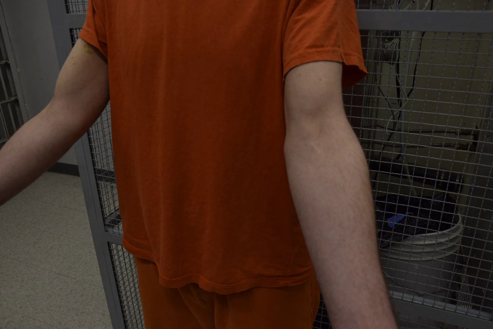 Torso in an orange shirt with both arms extended forward, against a metal mesh enclosure.