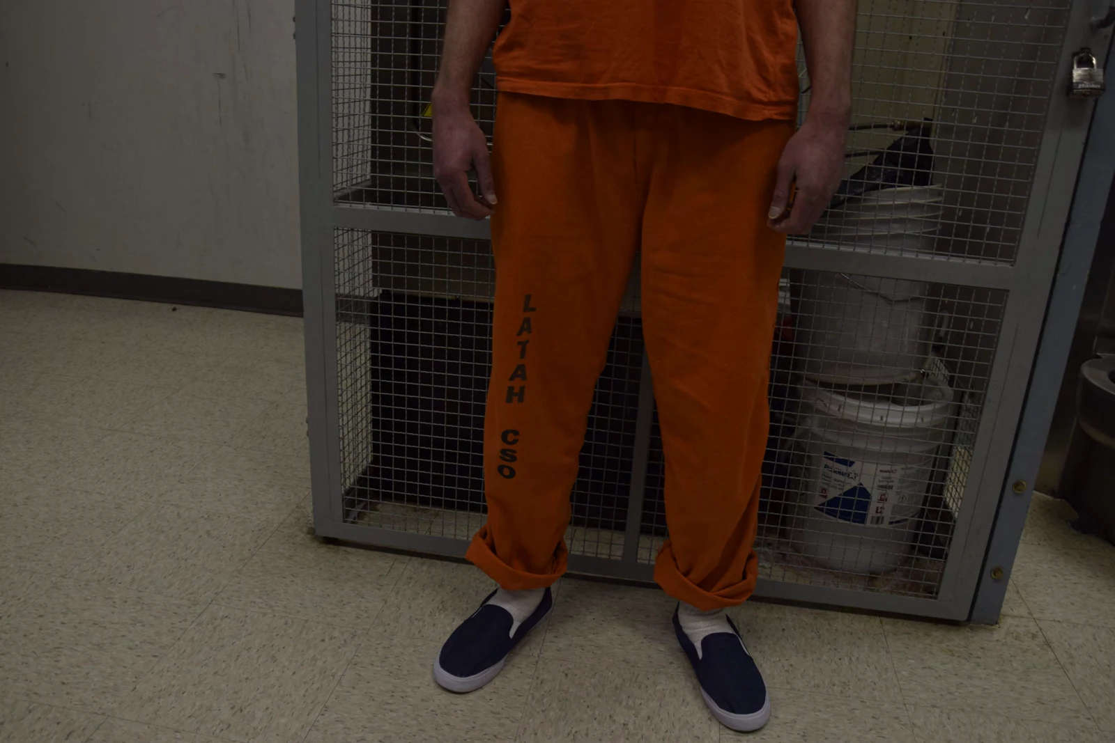 Lower-body view showing orange trousers labeled LATAH CO, blue slip-on shoes and hands resting at the sides.