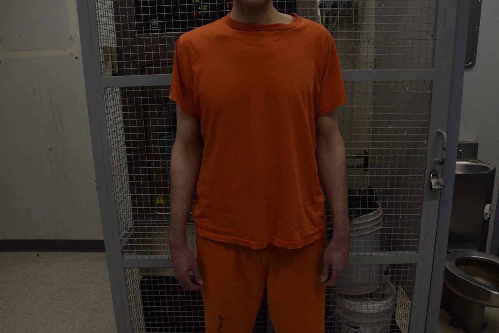 Frontal torso view of a person in orange clothing standing with arms at the sides in front of a metal mesh enclosure.