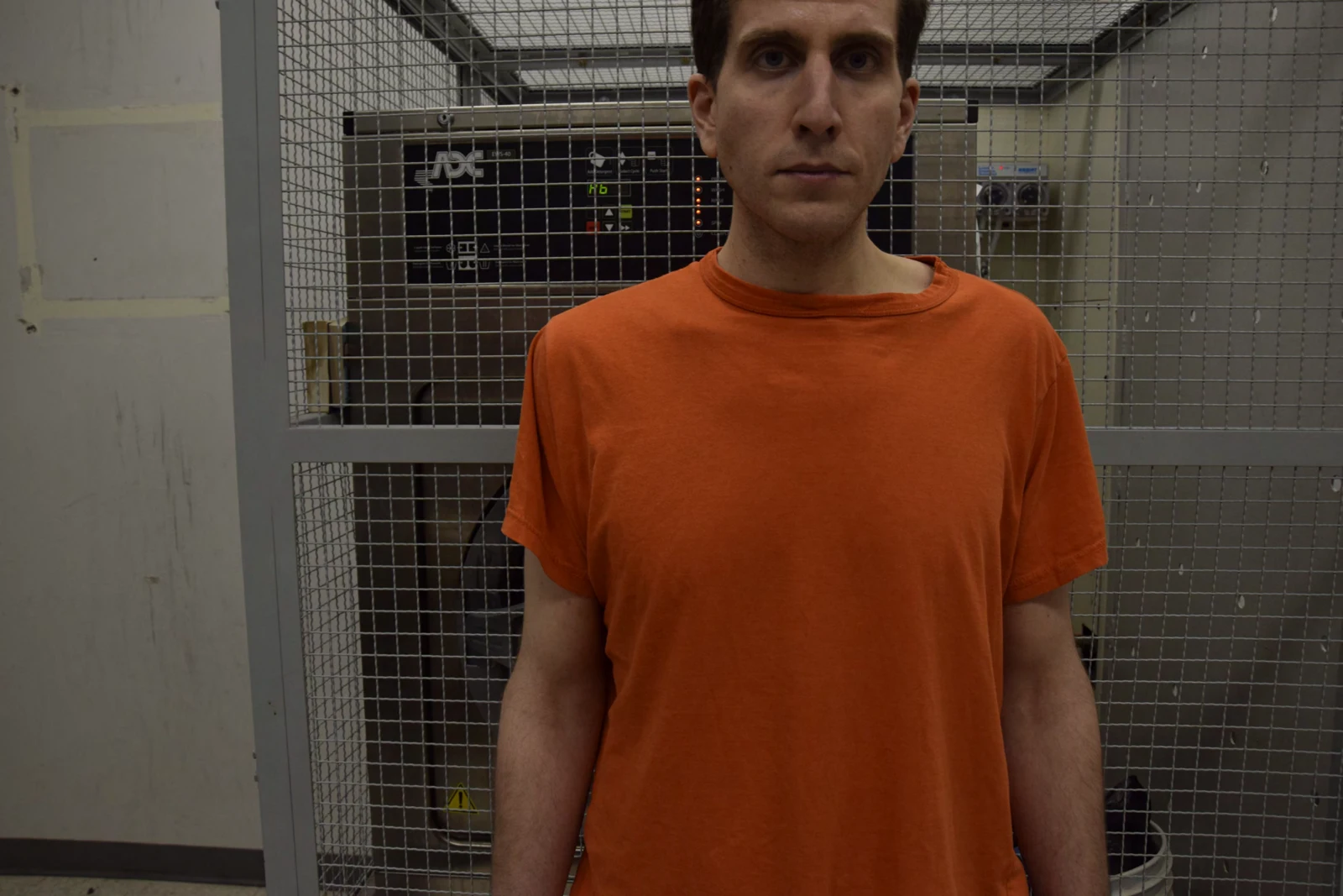 Front-facing upper-body portrait of a person in an orange shirt, against a mesh equipment enclosure.