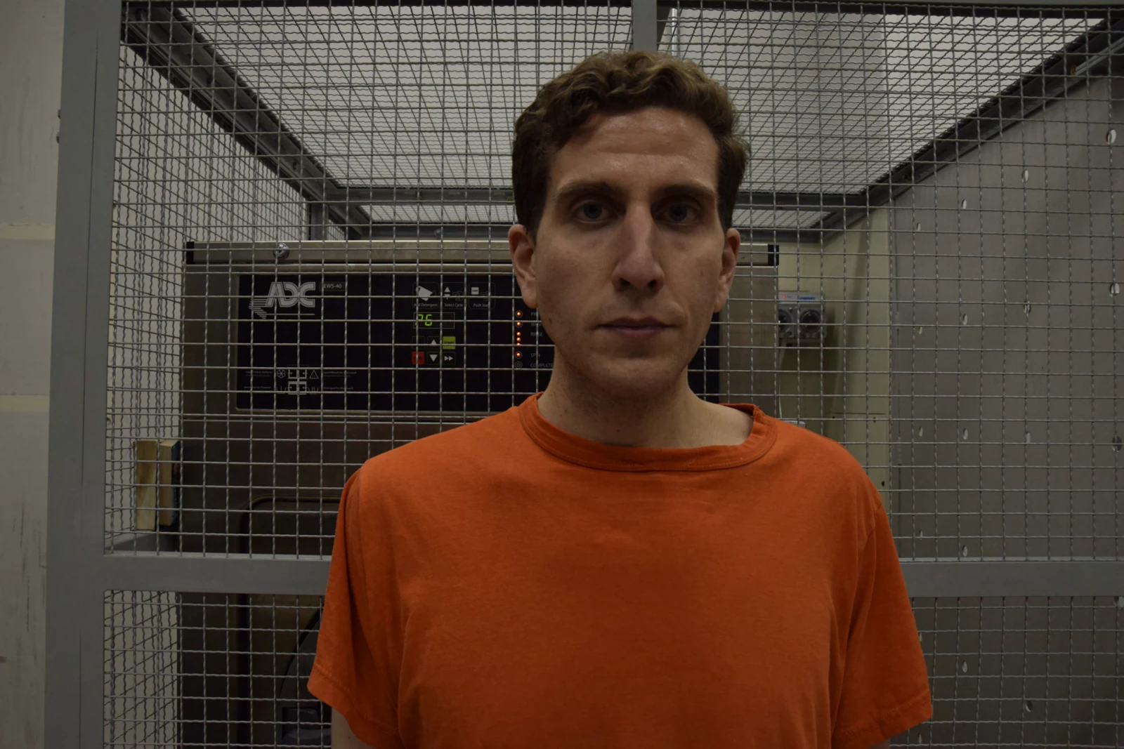 Closer front-facing head-and-shoulders portrait of a person with short dark hair in an orange shirt, against metal mesh.