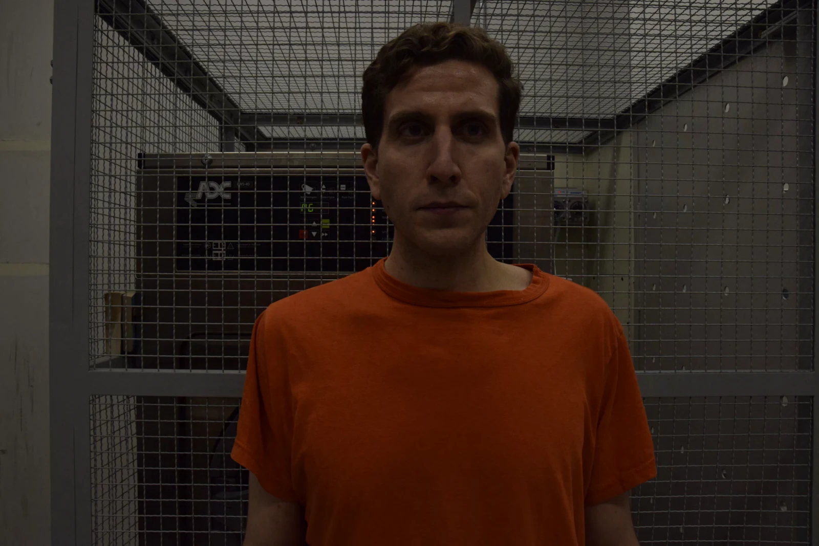 Front-facing head-and-torso portrait of a person in an orange shirt, against a metal mesh enclosure in dimmer lighting.
